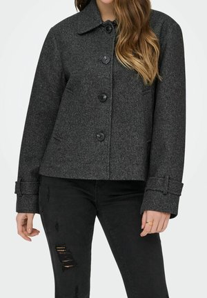 Femme portant un manteau en laine gris foncé boutonné avec de larges poignets, associé à un jean noir déchiré, debout sur un fond blanc uni.