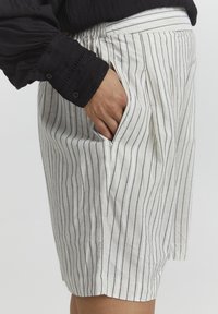 Shorts blancs rayés avec des lignes navy, fabriqués en tissu léger. Comprend des plis et des poches latérales, conçus pour le confort et la respirabilité.
