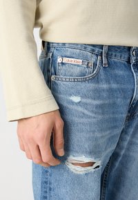 Ljusbeige långärmad topp i texturerat tyg, kombinerad med blå denimjeans med ett hål, ljus tvätt och Calvin Klein-märke på fickan.