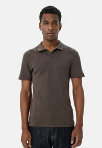 Brun ribbet polo skjorte med korte ærmer og klassisk krave, parret med mørkeblå jeans, båret af en person, der står foran en neutral baggrund.