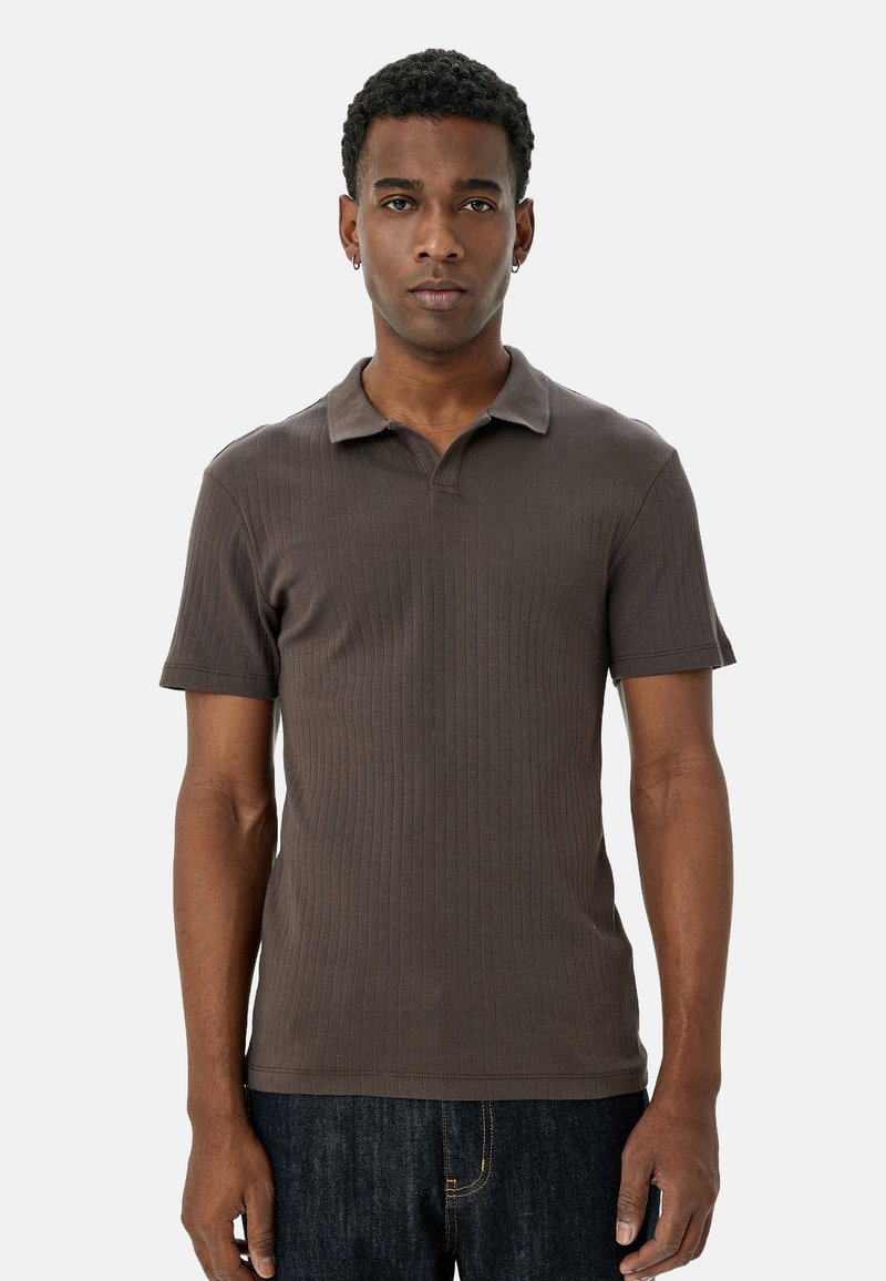 Brun ribbet polo skjorte med korte ærmer og klassisk krave, parret med mørkeblå jeans, båret af en person, der står foran en neutral baggrund.