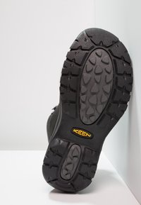 Suela de bota de senderismo negra con goma texturizada, que presenta un patrón de banda de rodadura multidireccional y un logo amarillo de "KEEN" para tracción.