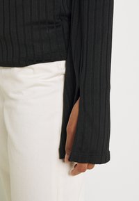 Schwarzes, geripptes Langarmoberteil mit seitlichem Schlitz, kombiniert mit weißen Jeans. Der Stoff wirkt weich und strukturiert.