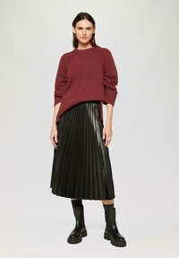 Pull en maille bordeaux avec des accents côtelés et des épaules tombantes, associé à une jupe plissée en simili cuir noire et des bottes noires imposantes.