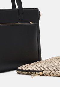 Sac à main en cuir noir avec poignées supérieures et pochette beige à motif avec fermeture éclair et tirette dorée, exposé sur une surface blanche.