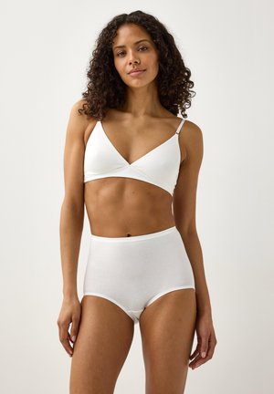 Donna con capelli ricci che indossa un bralette bianco e biancheria intima a vita alta, in piedi contro uno sfondo chiaro uniforme.