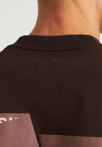 Bruine sweatshirt met een geribde halslijn, met een klein zwart lusdetail aan de achterkant en grote roze grafische accenten aan de zijkanten.
