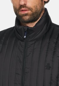 Schwarze isolierte Jacke mit vertikaler Naht, hohem Kragen, Frontreißverschluss und kleinem gestickten Pinguin-Logo auf der Brust. Glatte Textur.