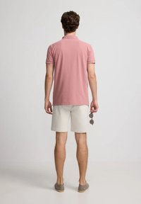 Camisa polo en rosa claro, de mangas cortas, con cuello acanalado. Combinada con pantalones cortos beige. Individuo sosteniendo gafas de sol en la mano izquierda.