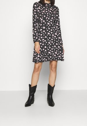 Robe noire à manches longues, ornée de motifs de cœurs roses et de pois blancs. Associée à des bottes de cowboy noires.