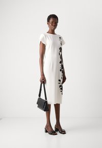 Abito fitted bianco con maniche corte e design floreale nero sul lato. La modella tiene una borsa nera e indossa sandali con tacco spesso neri.