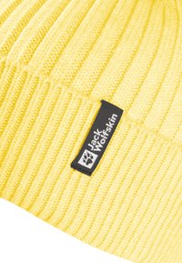 Gelber gerippter Strickstoff mit schwarzem rechteckigem Jack Wolfskin Logo-Tag, das einen weißen Pfotenabdruck und Text zeigt.