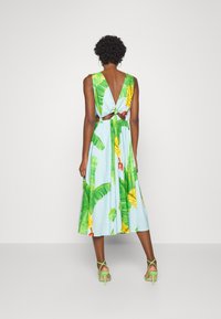 Blå midi-kjole med tropisk bladmønster og blomstermotiv, V-ryg design, knude detalje i taljen, parret med grønne hæle sandaler.