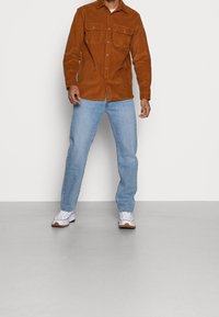 Camisa marrón de pana con botones y dos bolsillos en el pecho, combinada con vaqueros azul claro y zapatillas blancas con suelas marrones.