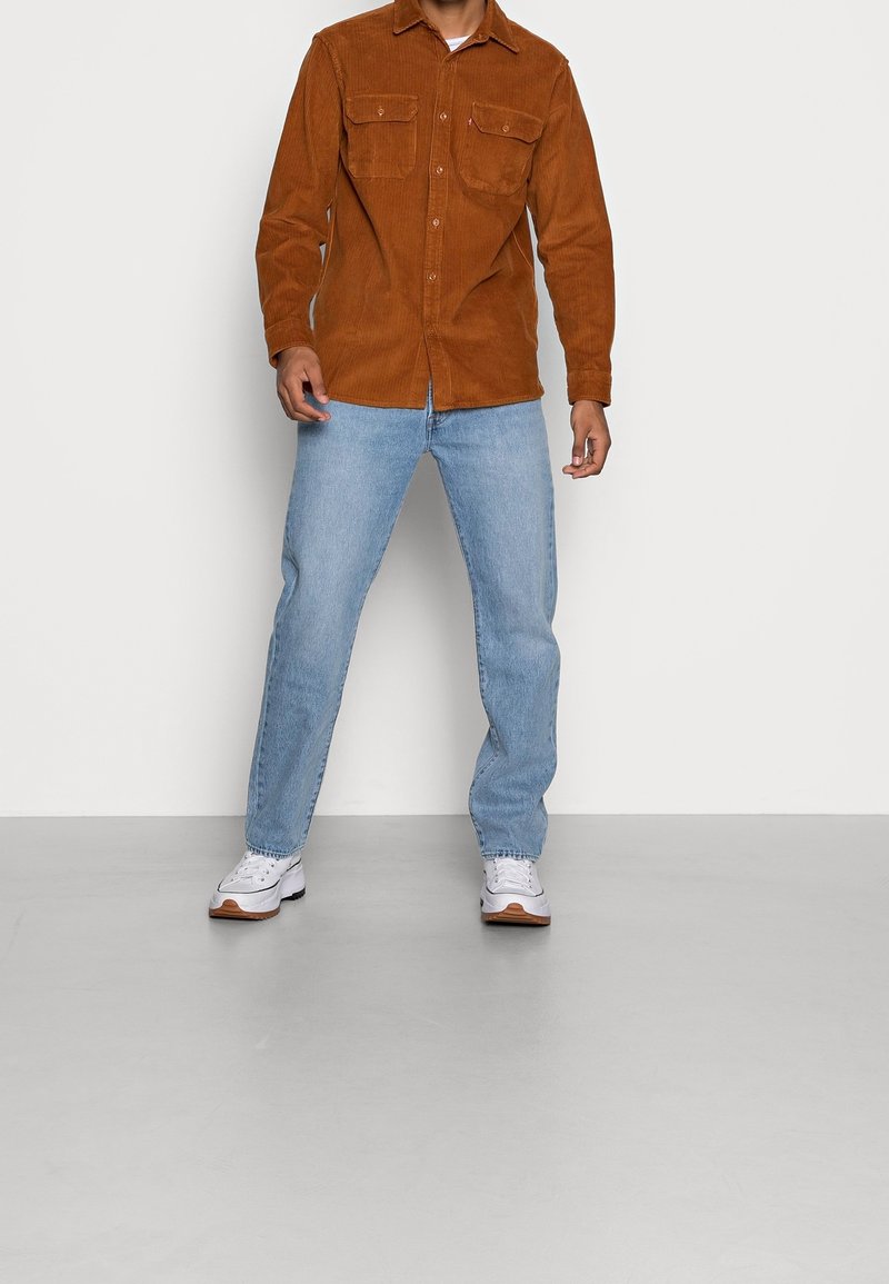 Camisa marrón de pana con botones y dos bolsillos en el pecho, combinada con vaqueros azul claro y zapatillas blancas con suelas marrones.