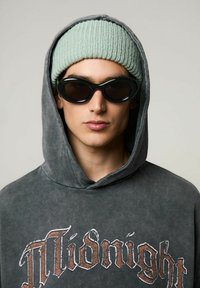 Felpa grigia oversize con stampa "Midnight", berretto a coste verde chiaro e occhiali da sole ovali neri. Primo piano del volto e degli accessori.