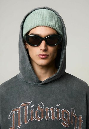Sudadera gris de gran tamaño con estampado de "Medianoche", gorro de punto acanalado verde claro, y gafas de sol ovaladas negras. Primer plano de la cara y los accesorios.