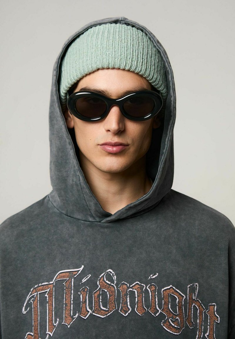 Felpa grigia oversize con stampa "Midnight", berretto a coste verde chiaro e occhiali da sole ovali neri. Primo piano del volto e degli accessori.