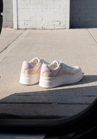 Paire de baskets compensées aux couleurs pastel posées sur un trottoir en béton devant un mur en briques gris clair.