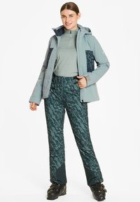 Lichtblauwe geïsoleerde jas met marineblauwe patroonaccenten, gecombineerd met slimfit teal camouflagebroek en zwarte skiboots.