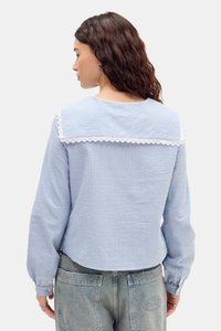 Lyseblå rutete bluse med hvit blondekantet krage og knappede mansjetter, laget av teksturert stoff. Brukt sammen med høyhalsede denimbukser.