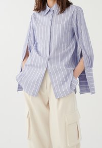 Chemise à rayures bleu clair et blanc avec col, manches longues ornées de boutons et une poche poitrine. Portée avec un pantalon beige.