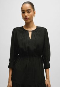 Frau mit glattem, nach hinten gezogenem Haar, die goldene Creolen und ein strukturiertes, schwarzes Kleid mit langen Ärmeln und einem Schlüsselaugen-Ausschnitt trägt.