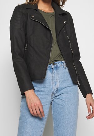 Zwarte leren motorjack met gewatteerde schouderaccenten, een gesneden kraag en zilveren ritsen, gestyled over een groene top en blauwe jeans.