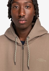 Beige zip-up hoodie featuring drawstrings with a green and yellow pattern and a small chest logo reading "ELEMENT SKATEBOARD CO. WORLDWIDE."