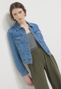 Giacca di jeans, azzurro chiaro, con due tasche frontali, chiusura a bottoni e polsini regolabili. Indossata sopra una maglietta grigia e pantaloni verde oliva.