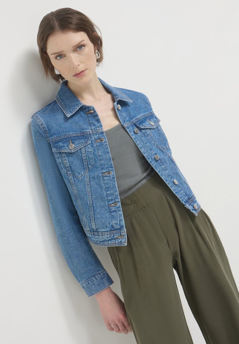 Giacca di jeans, azzurro chiaro, con due tasche frontali, chiusura a bottoni e polsini regolabili. Indossata sopra una maglietta grigia e pantaloni verde oliva.