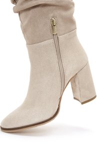 Bottine en suede beige à bout pointu, avec un talon épais et un détail de fermeture éclair sur le côté. Texture lisse et design simple.