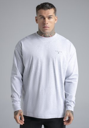 Bărbat cu tatuaje pe gât și pe mână, purtând un tricou cu mâneci lungi SikSilk de culoare gri deschis, stând în fața unui fundal simplu gri.