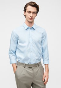 Νεαρός άνδρας με τα χέρια στις τσέπες, φοράει ένα ανοιχτόγαλανό πουκάμισο με κουμπιά και ρεβέρ στα μανίκια και μπεζ παντελόνι, μπροστά σε απλό φόντο.