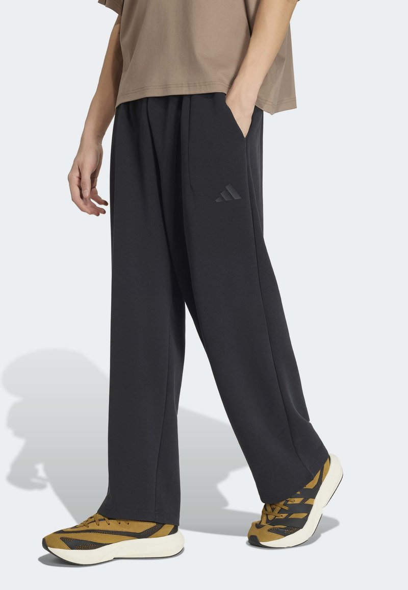 Personne portant un pantalon Adidas noir ample avec la main dans la poche et des baskets marron et noires, debout sur un sol gris clair.