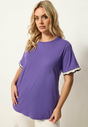 Blonde Frau trägt ein locker sitzendes lila Kurzarmtop mit weißem und schwarzem wellenförmigem Besatz an den Ärmeln, steht vor einem schlichten Hintergrund.