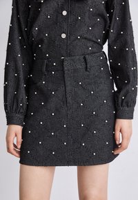 Jupe en denim noir avec motif matelassé, ornée de perles blanches. Elle comprend une ceinture et des poches avant. Texture lisse.
