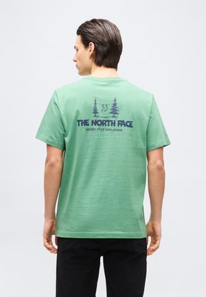 Man met een groen T-shirt met het logo van "The North Face", bomen en een afbeelding van wandelschoenen op de achterkant, gecombineerd met een zwarte broek.