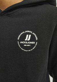 Črna kapuca z okroglo belo oznako. Besedilo vključuje "RESEARCH_DESIGN_APPAREL" in "JACK&JONES ESS. DEPT." v poudarjenem tisku. Gladka tekstura.
