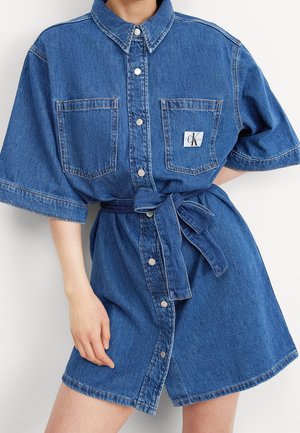 Robe en denim bleu à manches courtes avec fermeture boutonnée devant, deux poches poitrine, une ceinture nouée à la taille et un patch logo sur la poche gauche.