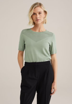 Haut vert clair à manches courtes en tissu doux avec un détail de motif sur l'épaule. Porté avec un pantalon noir taille haute.