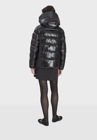 Svart vadderad jacka med glänsande yta, utrustad med stor huva, horisontellt quiltad design och ribbade muddar, bärs över en svart kjol.