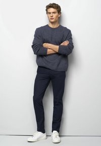Pull navy, tricot texturé avec des poignets côtelés, associé à un pantalon navy ajusté et des baskets blanches. Le modèle pose avec les bras croisés devant un fond uni.
