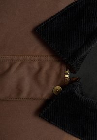 Brunt stof med teksturdetaljer, der har en sort corduroy krave og en metal snapknap. Lynlåsen er synlig nær kraveområdet.