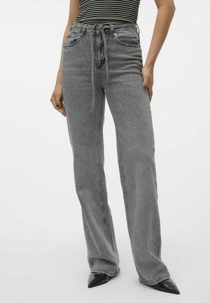 Vrouw draagt grijze high-waist rechte jeans met een geknoopte stoffen riem, zwarte puntige schoenen en een zwart-wit gestreepte mouwloze top.
