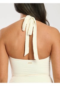 Vit texturerad halterneck-top med knytband i nacken, med ett subtilt mönster och en liten logotags på baksidan, som visar en smidig silhuett.