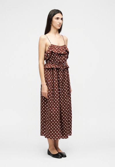 Γυναίκα που φοράει καφέ μίντι φόρεμα με λευκά πουά, με βολάν στα τιράντες και ζώνη στη μέση, φοράει μαύρα φλατ παπούτσια σε μονόχρωμο φόντο.