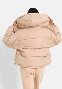 Vadderad, beige dunjacka med hög krage, synliga sömmar och en avslappnad passform, bärs över matchande beige byxor.
