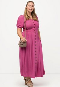 Fuchsiové lněné šaty s nabíranými rukávy a knoflíkovou přední částí, zdobené knoflíky ve dřevěné barvě. Modelka nosí malou kabelku přes rameno a sandály na podpatku.