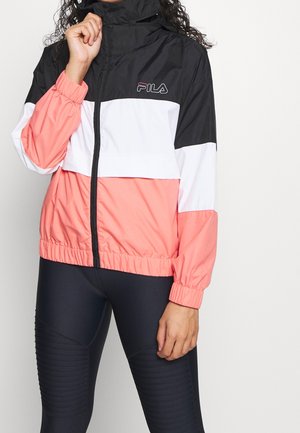 Woman wearing a black, white, and coral Fila windbreaker jacket with elastic cuffs and black textured leggings.